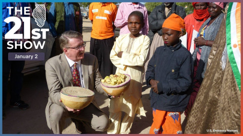 a white man in a tan suit crouches down and presents two gourds to a pair of Black children in traditional Nigerien dress — a boy wearing a long, cream-colored shirt and a girl in an orange dress with blue flowers embroidered on it; 