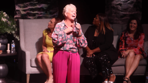 Woman stands on stage with microphone, other women sit behind her
