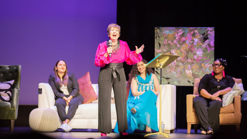 Woman holding a mic on stage laughing, women sit on couches behind her