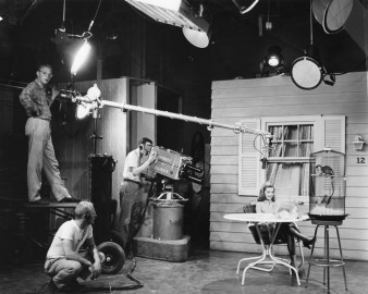 woman sits on set with monkey, one man operates camera, one man operated mic boom, another squats