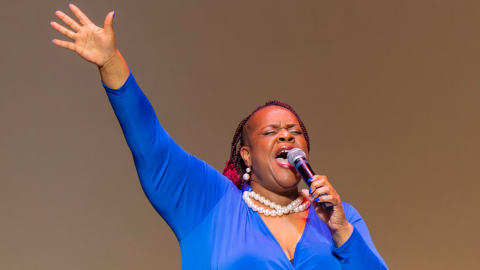 A woman in blue top and pearls talks on microphone with hand up