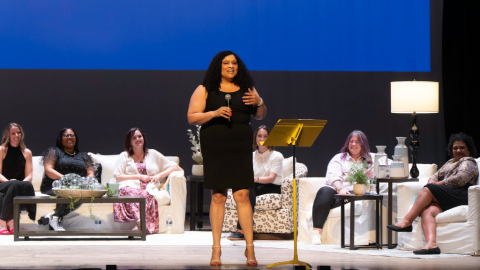woman in black dress holds microphone on stage with others sitting on couches behind her