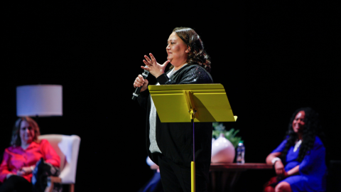 Woman stands on stage with microphone, other women sit behind her