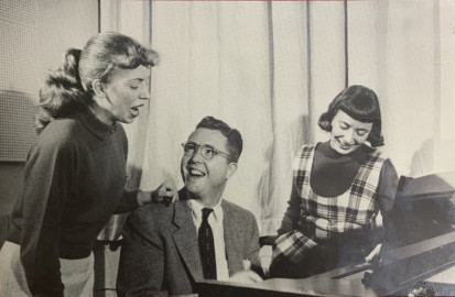 man sits at piano with a woman standing on other side, man smiles, woman sings