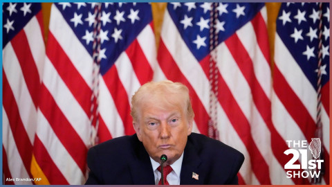 Donald Trump sitting before a microphone, with a wall of American flags behind him