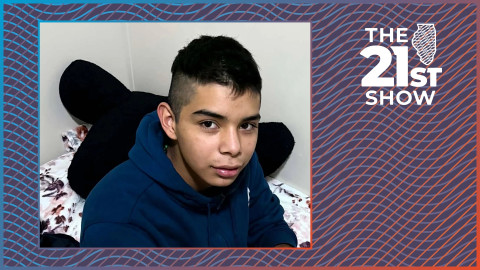 a teenager with dark hair wearing a navy blue hoodie is sitting on a bed