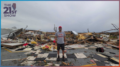 a white man with with a beard in a red hat, gray T-short, and dark shorts; he is standing on the broken pieces of a black-and-white checkered floor amid the debris of a building, including pieces of wood, bits of insulation, and piles of twisted corrugated metal; the trunk and a few freshly stripped limbs of a tree are in the background