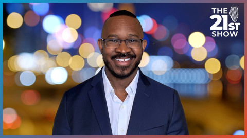 a Black man wearing a navy suit and light-colored shirt with an open collar stands in front of a blurred cityscape