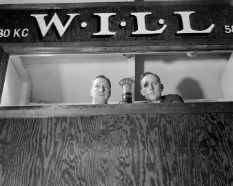 Two men sit behind microphones in WILL Huff Gym broadcasting booth in 1944