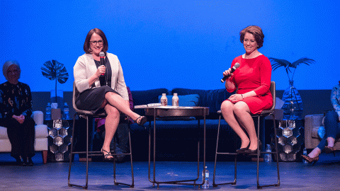 two women sit on barstools on a stage