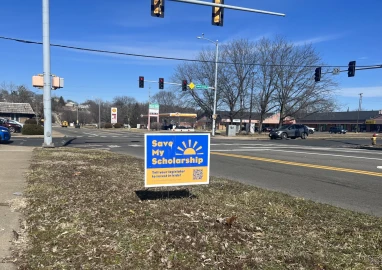 A "Save My Scholarship" sign sits at the intersection of John H. Gwynn Jr Ave. and MacArthur Highway in Peoria.