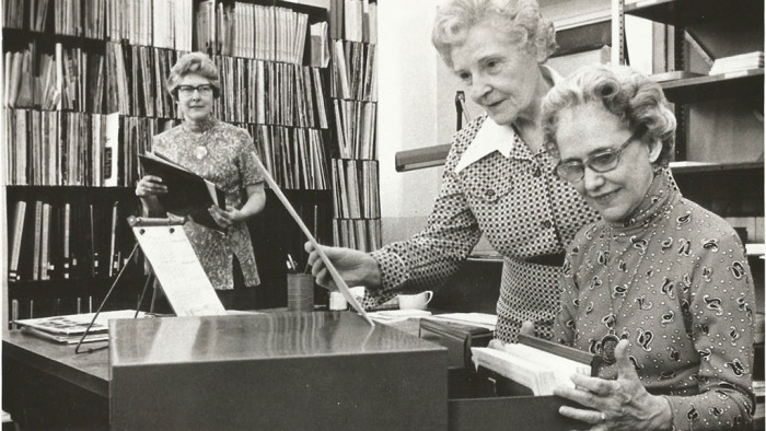 three women stand in room looking in filing cabinet